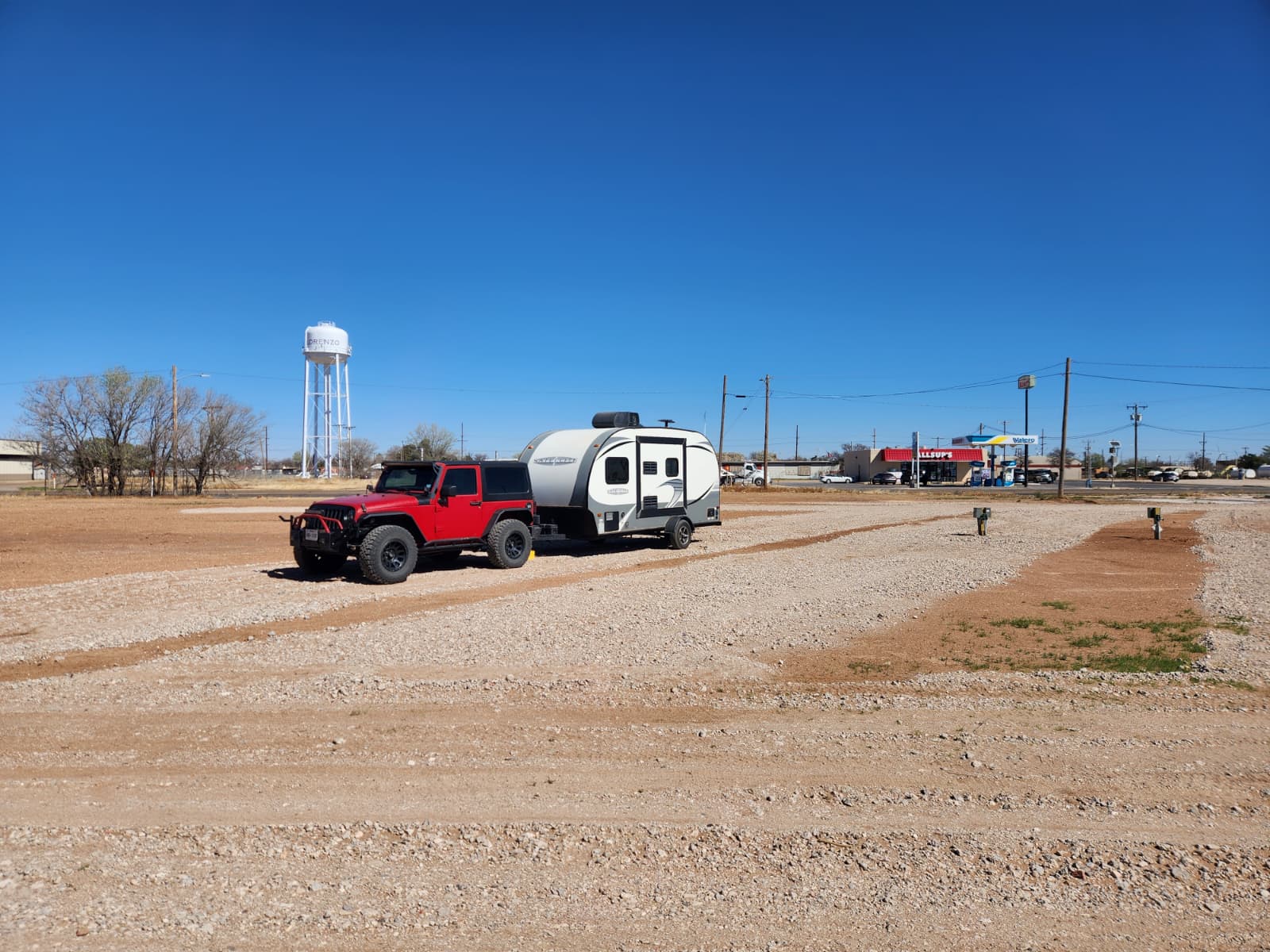 A red Jeep towing a small white travel trailer is parked on a gravel lot under a clear blue sky, with a water tower and buildings visible in the background.