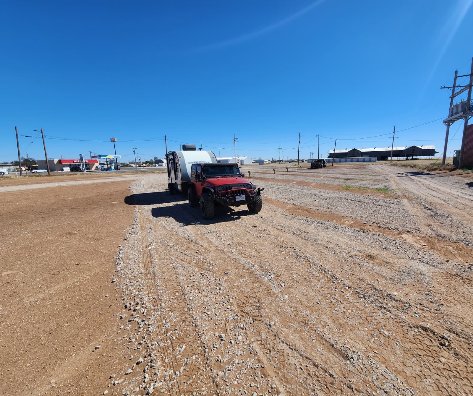 A red off-road vehicle is towing a small white trailer in a large, empty, gravel-covered lot under a clear blue sky. Some buildings and utility poles are visible in the background.