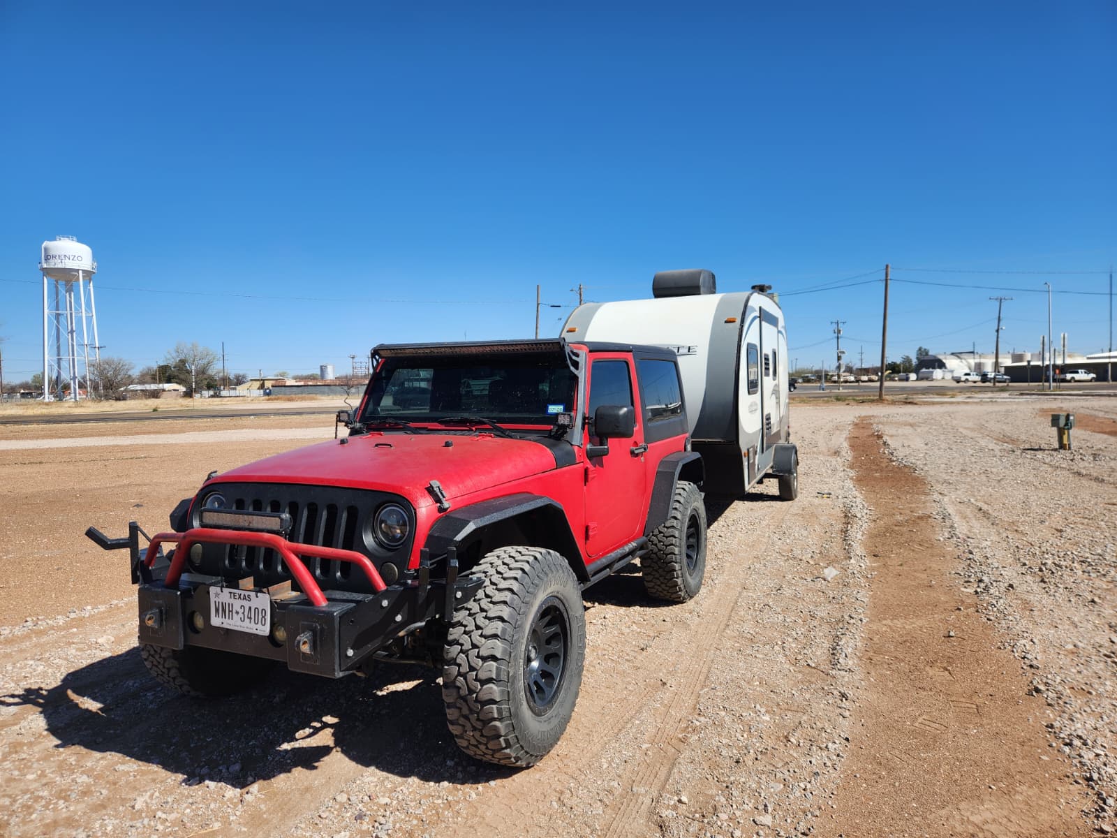 A red Jeep with off-road tires is towing a small white travel trailer on a dirt road. A water tower and power lines are visible in the background under a clear blue sky.