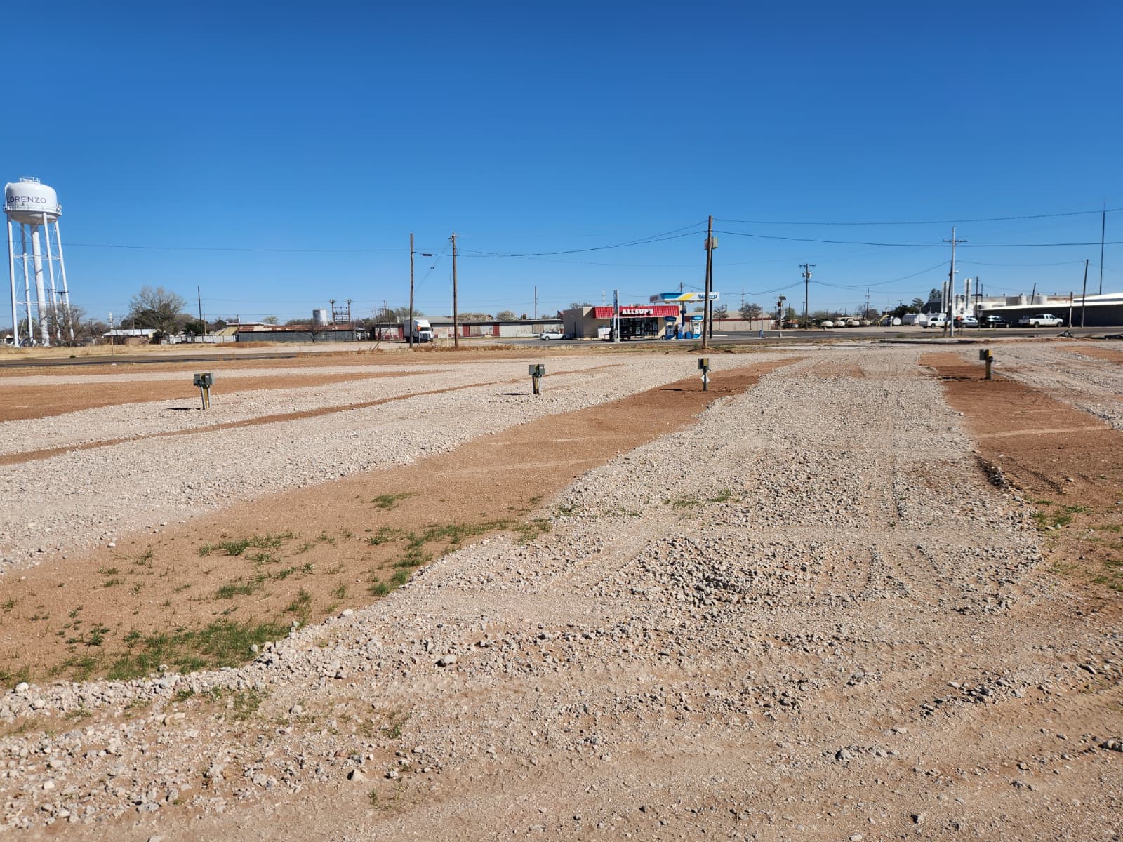 A gravel lot with RV hookups, sparse grass patches, utility connections, and a water tower and buildings visible in the background under a clear blue sky.