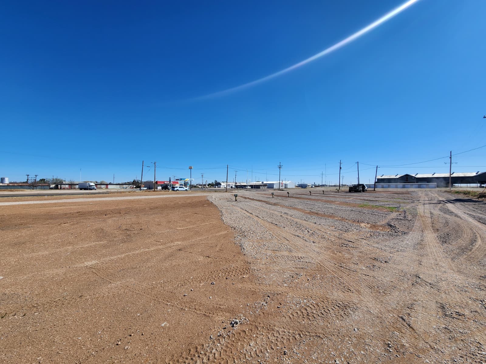 A wide, open dirt lot with tire tracks under a clear blue sky, bordered by buildings, utility poles, and a few vehicles in the background.